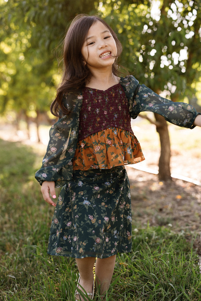 A young girl with long dark hair smiles outdoors, standing on grass with trees in the background, wearing the Ivy City Co Mini August Set in Elysian Vine—a floral dress featuring a smocked bodice with maroon and orange patterns, balloon sleeves, and a green floral skirt.