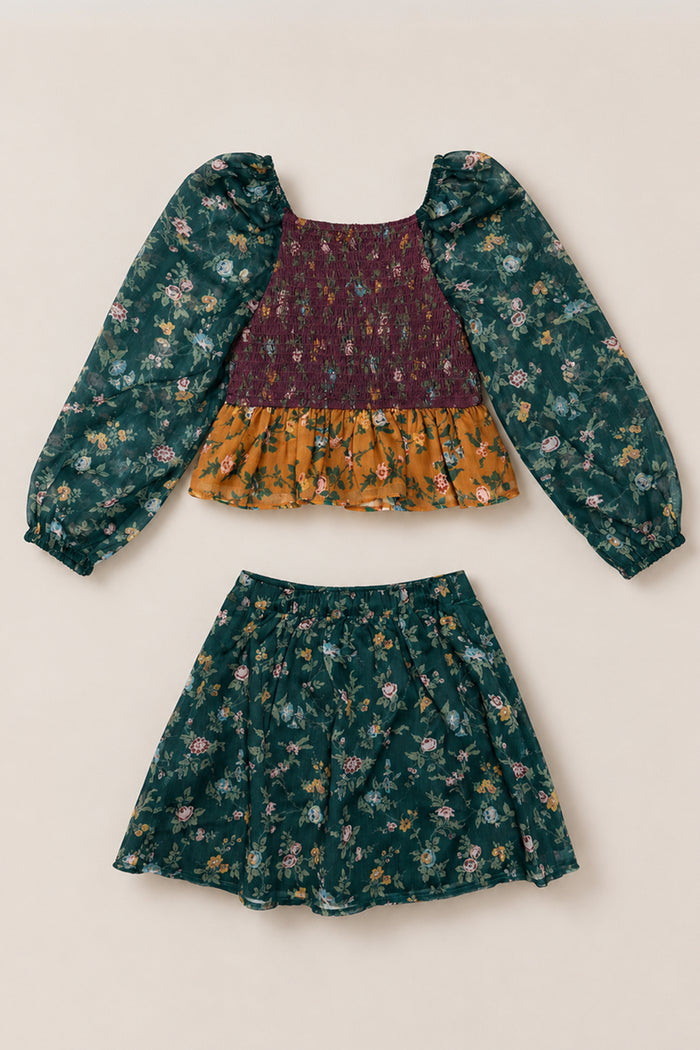 The Mini August Set in Elysian Vine by Ivy City Co is a two-piece ensemble featuring a floral-patterned green long-sleeve top with an elegantly smocked bodice, subtle balloon sleeves, and a mustard ruffle accent. Paired with a coordinating green floral skirt, both pieces are artfully displayed flat against a beige background.