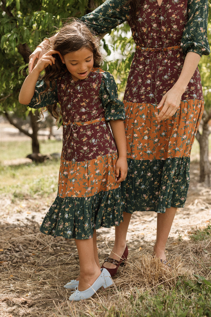 An adult woman and a young girl stand outdoors amid grass and trees, sunlight streaming through the branches. Both are dressed in matching Ivy City Co Mini Harlowe Dresses in Elysian Vine, featuring charming puff sleeves and crafted from soft viscose fabric. The woman gently touches the girl’s hair as they share a tender moment.