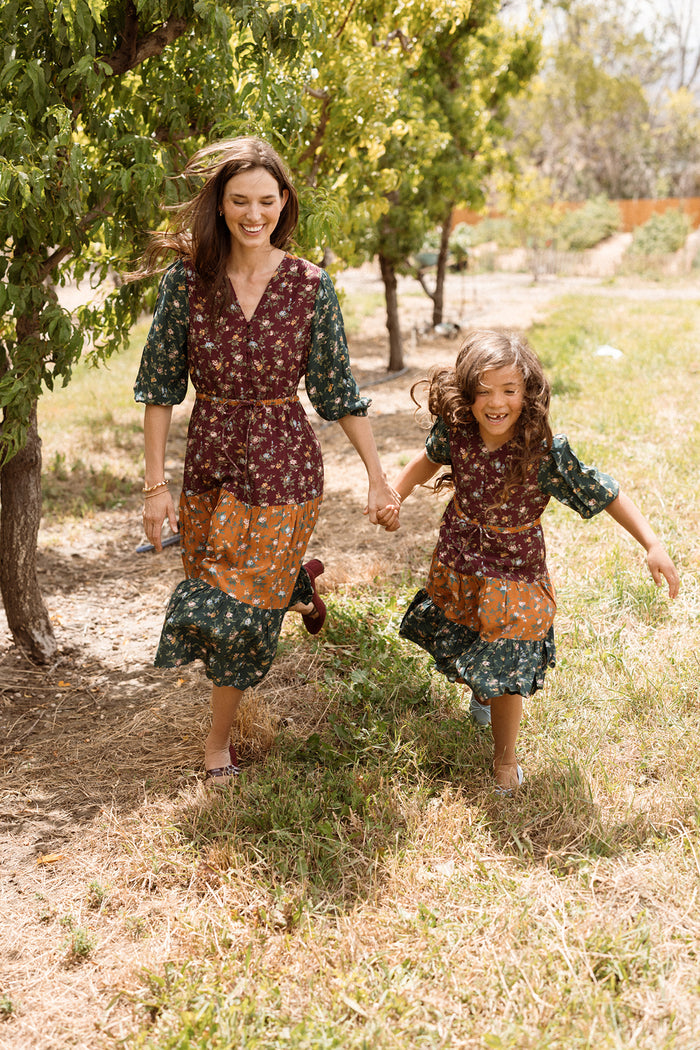 A woman and a young girl, both wearing matching Ivy City Co’s Mini Harlowe Dress in Elysian Vine—featuring floral viscose fabric and charming puff sleeves—hold hands and smile as they walk along a tree-lined grassy path outdoors.