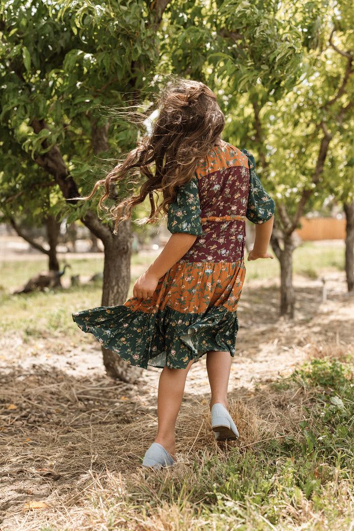 A young girl with long, wavy hair wearing the Ivy City Co Mini Harlowe Dress in Elysian Vine with puff sleeves walks away through an orchard, her hair and dress flowing in the wind.