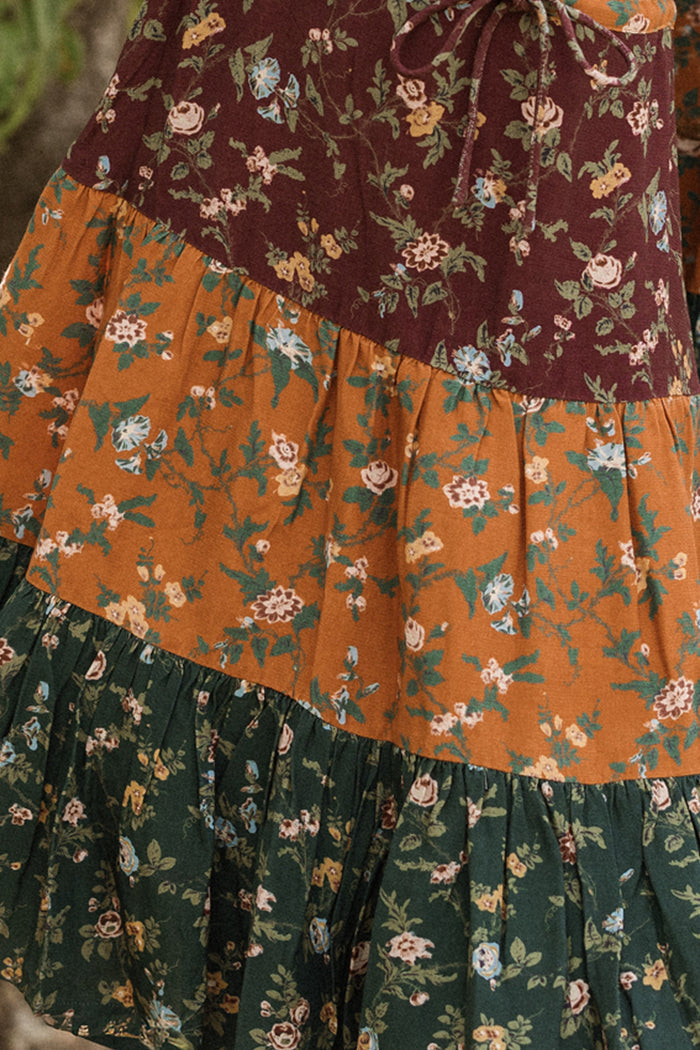 A close-up of the Mini Harlowe Dress in Elysian Vine by Ivy City Co showcases its tiered silhouette, crafted from soft viscose with sections of maroon, orange, and dark green floral fabric—each patterned with petite flowers and leafy details. Delicate puff sleeves add a charming finishing touch.