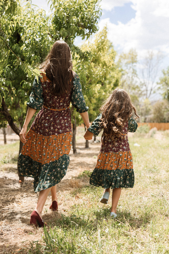 A woman and a young girl, both wearing the charming Ivy City Co Mini Harlowe Dress in Elysian Vine with delightful puff sleeves and matching patterned viscose fabric, hold hands as they walk outdoors along a sunlit tree-lined path.