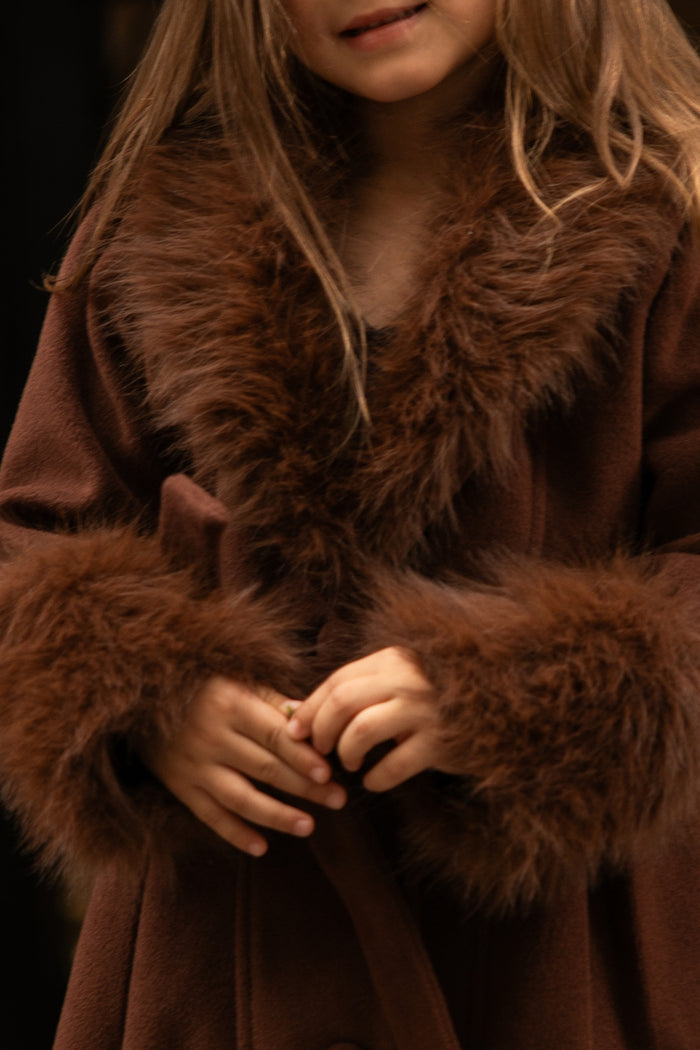A young girl with blonde hair wears the Ivy City Co Mini Pookie Coat in Warm Cocoa, featuring a cozy design with fluffy fur trim at the collar and cuffs, her hands clasped together.