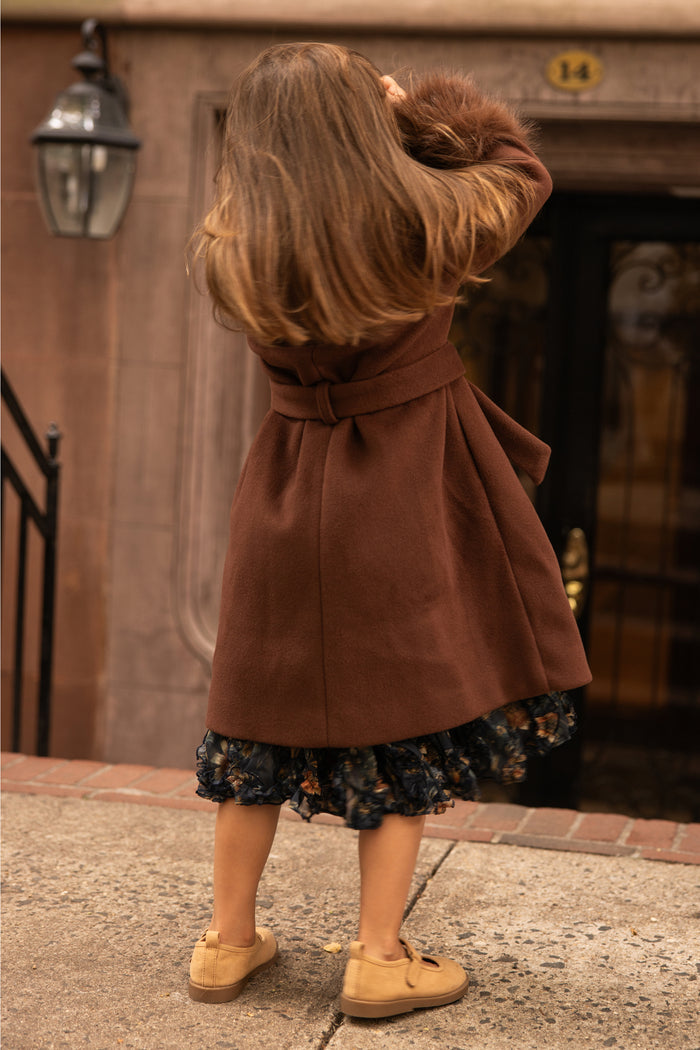A person with long hair is wearing the Ivy City Co Mini Pookie Coat in Warm Cocoa—a girls’ fall coat—paired with a patterned skirt and tan shoes, standing on a sidewalk in front of a brownstone building with a black door and the number 14 above it.