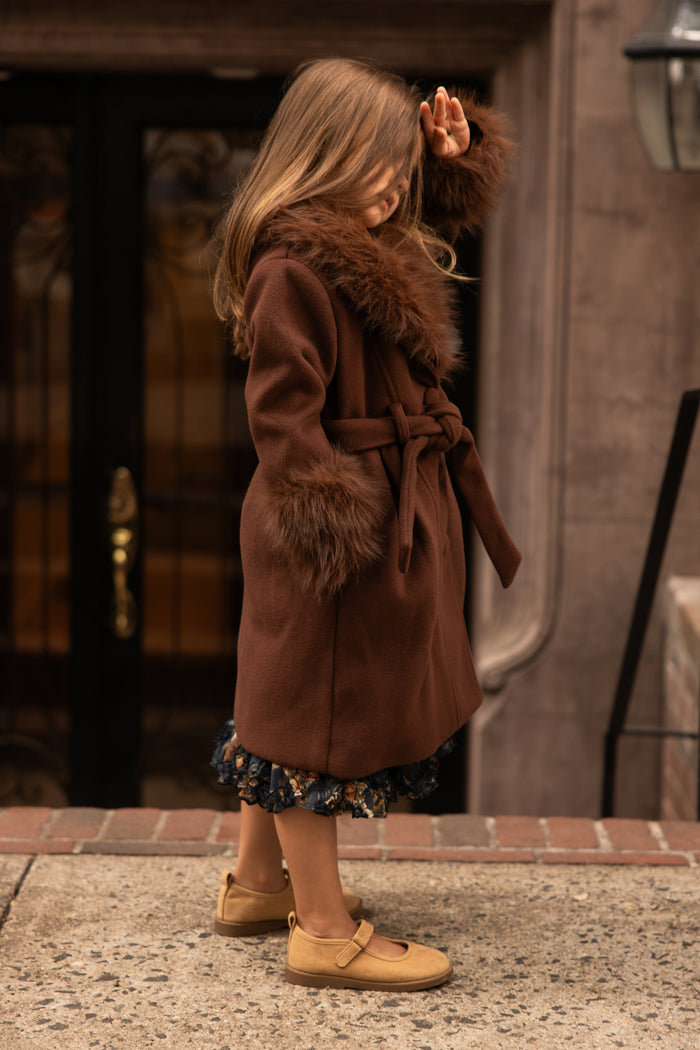 A young girl wearing the Ivy City Co Mini Pookie Coat in Warm Cocoa with fur trim and tan shoes stands on a sidewalk, partially shielding her face with her hand, in front of a building entrance.