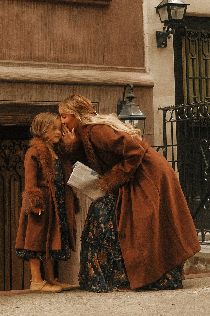 A woman whispers to a smiling girl on a city sidewalk by a brownstone building. Both wear brown coats with fur trim and floral dresses. The girl’s coat, the Mini Pookie Coat in Warm Cocoa by Ivy City Co, blends comfort with classic autumn style as the woman holds a newspaper.