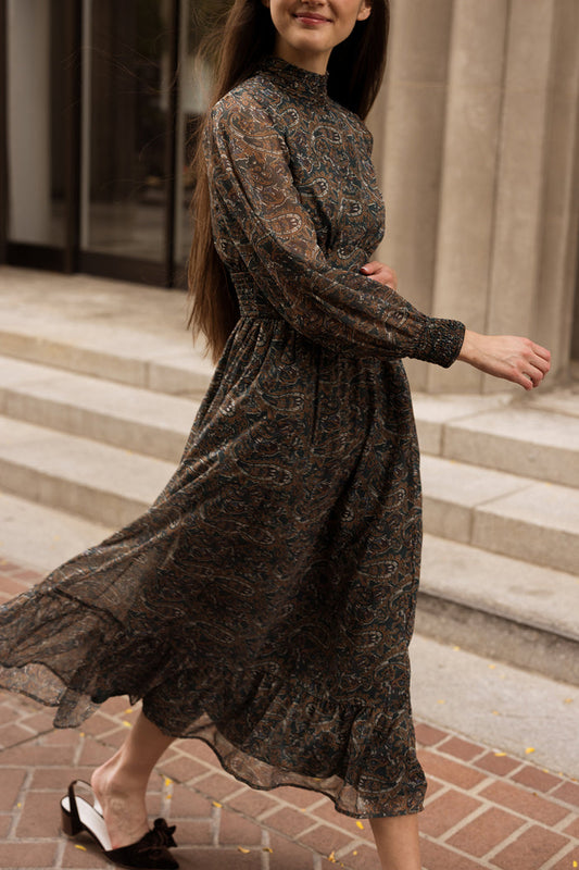 A woman in the Ivy City Co Payton Dress in Evergreen Paisley—a long, patterned sheer maxi dress with long sleeves—walks outside near stone steps. She has long brown hair and is wearing black slip-on shoes with bows. Her face is partly visible, and she smiles slightly.