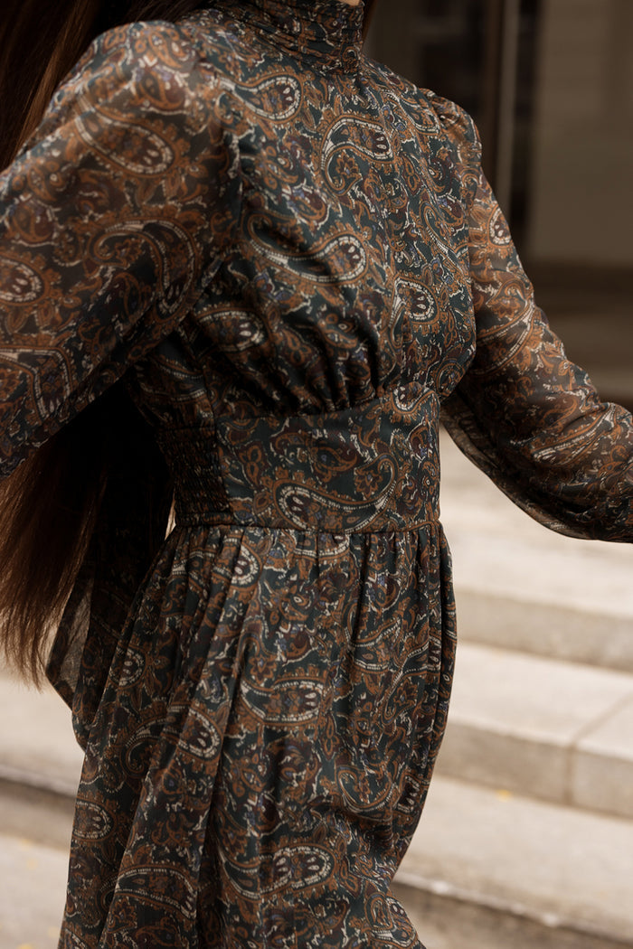 A person wearing the Ivy City Co Payton Dress in Evergreen Paisley—a long-sleeved maxi dress with a high neckline and a brown and green paisley pattern—is walking outdoors, with stone steps visible in the background.