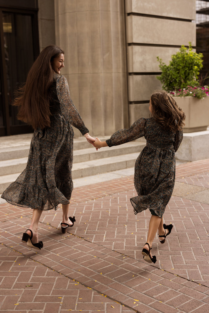 A woman and a girl, both wearing the Ivy City Co Payton Dress in Evergreen Paisley with heels, hold hands and walk joyfully along a brick sidewalk near a building adorned with large columns and flowers.