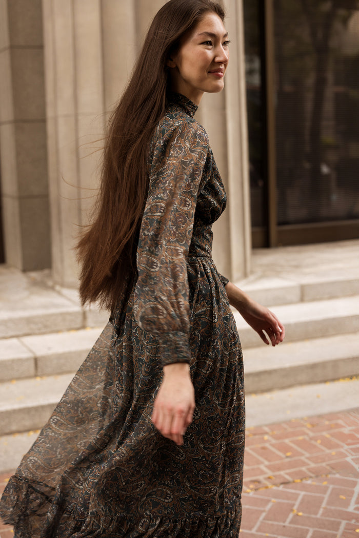 A woman with long brown hair wears the Ivy City Co Payton Dress in Evergreen Paisley as she walks outdoors on a brick sidewalk near stone steps and tall buildings.