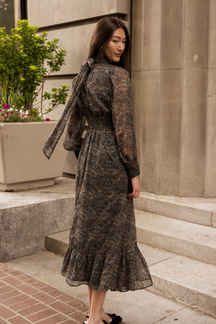 A woman wearing the Ivy City Co Payton Dress in Evergreen Paisley stands near stone steps outdoors, glancing over her shoulder. She pairs the long-sleeved maxi dress with black shoes, while a large planter filled with greenery and flowers sits nearby.
