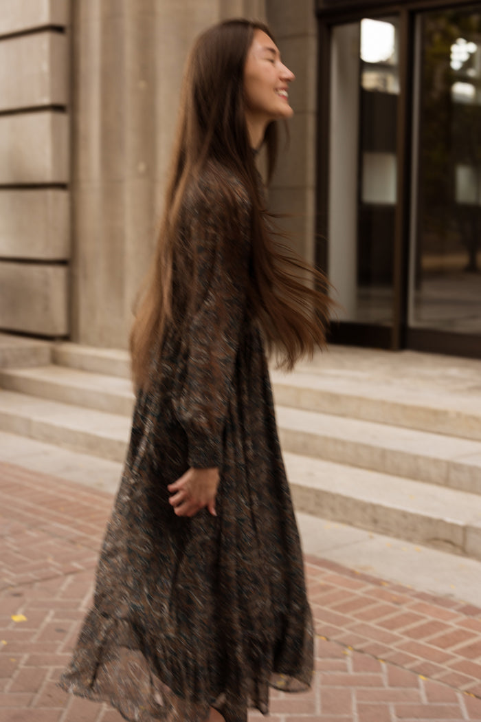 A woman with long brown hair walks and smiles outdoors in front of stone steps and large glass doors, wearing the Ivy City Co Payton Dress in Evergreen Paisley—a flowy maxi dress. The softly blurred image conveys a sense of movement.