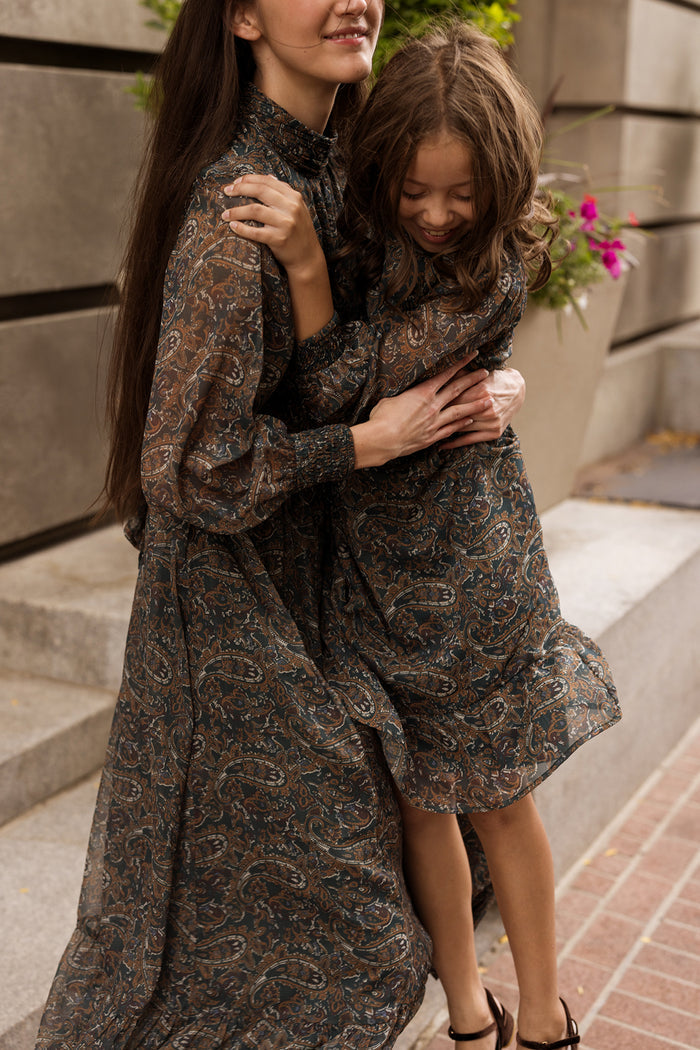 An adult woman and a young girl, both wearing the Ivy City Co Payton Dress in Evergreen Paisley, smile and embrace while sitting together on outdoor concrete steps near a flower planter.