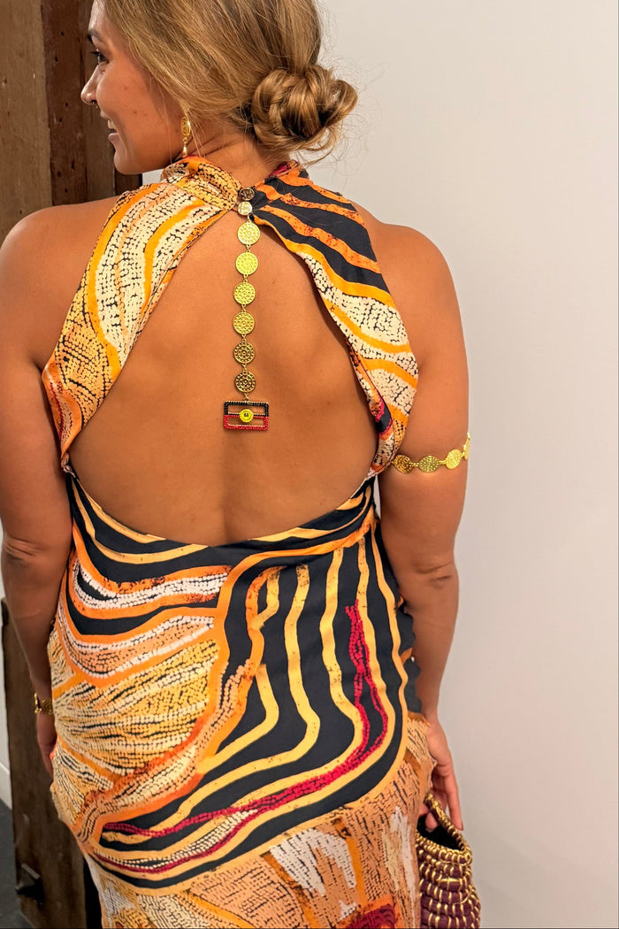Aboriginal Woman in a First Nations designed dress with a open back and Aboriginal flag jewellery  standing against a white wall.