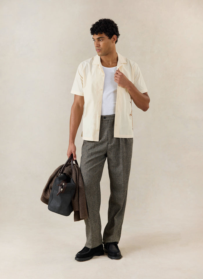 A model wears the cream Percival Supper Club Embroidered Cuban Shirt over a white vest, with grey trousers and a holdall.