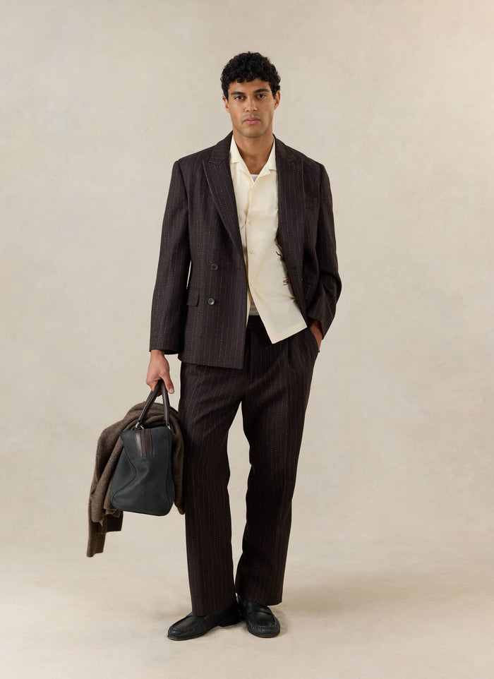 A model wears the cream Percival Cuban shirt under a brown pinstripe suit, holding a dark bag and jacket, with black loafers.