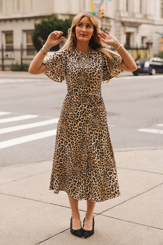 A woman wearing the Tallulah Dress in Leopard by Ivy City Co stands on a city sidewalk, raising her hands near her head. She has long blonde hair and is wearing black heels. Buildings, a crosswalk, and a parked car are visible in the background.