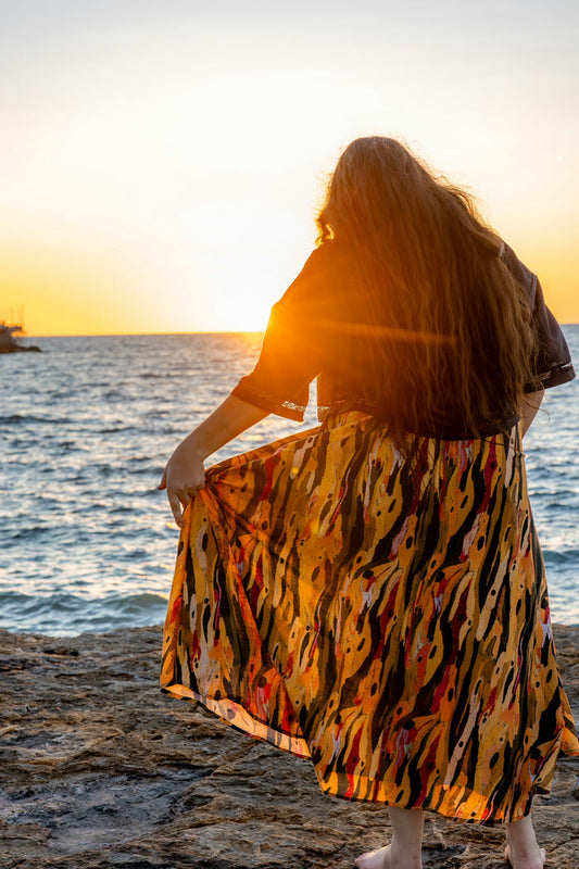 Woman in a colorful Aboriginal artwork skirt standing on a beach at sunset.