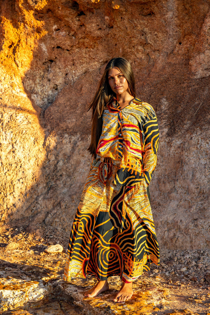 Woman in a colorful Aboriginal artwork Long sleeved top and skirt, standing against a rocky wall