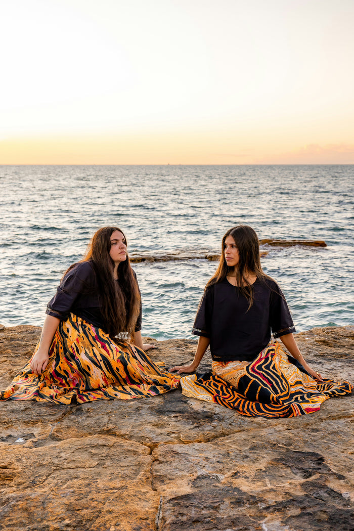 Two Aboriginal women sitting on rocks wearing Aboriginal artwork skirts by the ocean at sunset.